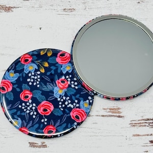 May include: A compact mirror with a floral design. The mirror has a blue background with pink and white flowers. The mirror is closed and the floral side is facing up.