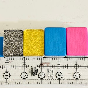 May include: Five small square tiles with different patterns and colors. The tiles are lined up in a row from left to right: light blue, silver glitter, gold glitter, light blue with a geometric pattern, and pink with a geometric pattern. The tiles are sitting on a white surface with a ruler underneath.
