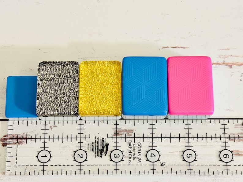 May include: Five small square tiles with different patterns and colors. The tiles are lined up in a row from left to right: light blue, silver glitter, gold glitter, light blue with a geometric pattern, and pink with a geometric pattern. The tiles are sitting on a white surface with a ruler underneath.