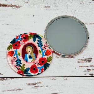 May include: A round, open compact mirror with a floral design and a portrait of a girl in a blue dress. The mirror is open, revealing the reflective surface. The floral design includes red poppies and blue leaves.