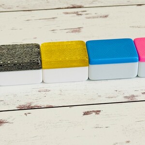 May include: Five small square boxes with white bases and different colored tops. The boxes are arranged in a row from left to right. The colors are blue, black and white, gold, blue, and pink.