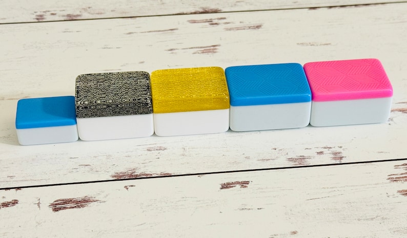 May include: Five small square boxes with white bases and different colored tops. The boxes are arranged in a row from left to right. The colors are blue, black and white, gold, blue, and pink.