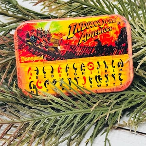 Può includere: Uno spillo in metallo rosso, giallo e nero con le parole "Indiana Jones Adventure" e "Disneyland" su di esso. Lo spillo presenta una scena del giro di Indiana Jones Adventure a Disneyland.