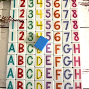 May include: A set of colourful tiles with numbers 1 to 8 and letters A to H. The tiles are arranged in a grid pattern on a white wooden surface. A blue tile is placed in the centre of the grid.
