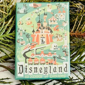 Può includere: Un magnete verde e bianco con un'illustrazione in stile vintage di Disneyland. Il magnete include il testo "Disneyland 60 Years of Dreams".