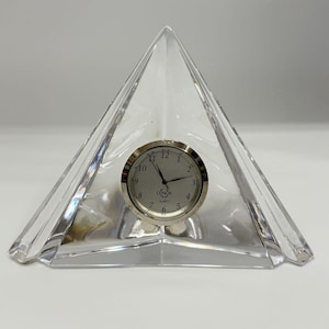 May include: A clear, triangular glass desk clock featuring a silver-toned circular face with black Arabic numerals and hands. The clock face is centered within the glass structure and displays the brand name Lenox and the word Quartz.