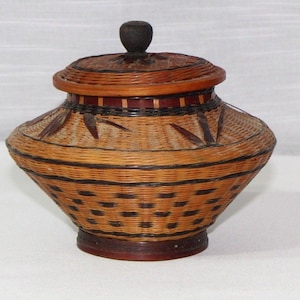 May include: A handcrafted, lidded basket with a woven design. The basket features a round, dark wooden knob on top of the lid. The body of the basket is a light brown color with dark brown accents and a decorative pattern.