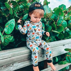 May include: A child wearing a gray pajama set with a colorful pattern of hearts, skulls, and other graphics, paired with a black baseball cap. The child is sitting on a wooden fence, with green foliage in the background.