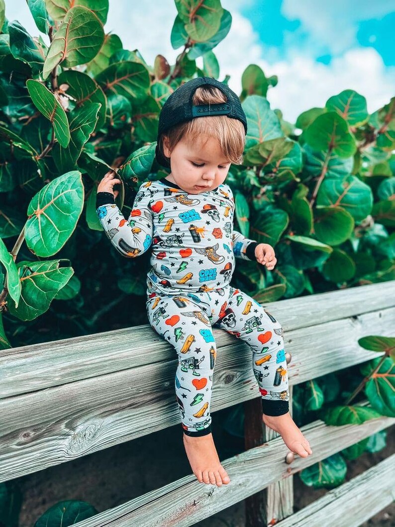 May include: A child wearing a gray pajama set with a colorful pattern of hearts, skulls, and other graphics, paired with a black baseball cap. The child is sitting on a wooden fence, with green foliage in the background.