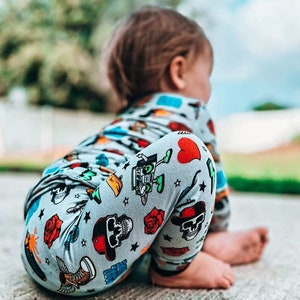 May include: A child wearing a light gray onesie and leggings with a repeating pattern of skulls, roses, hearts, and other graphics. The child is sitting on a concrete surface, with their back to the camera.