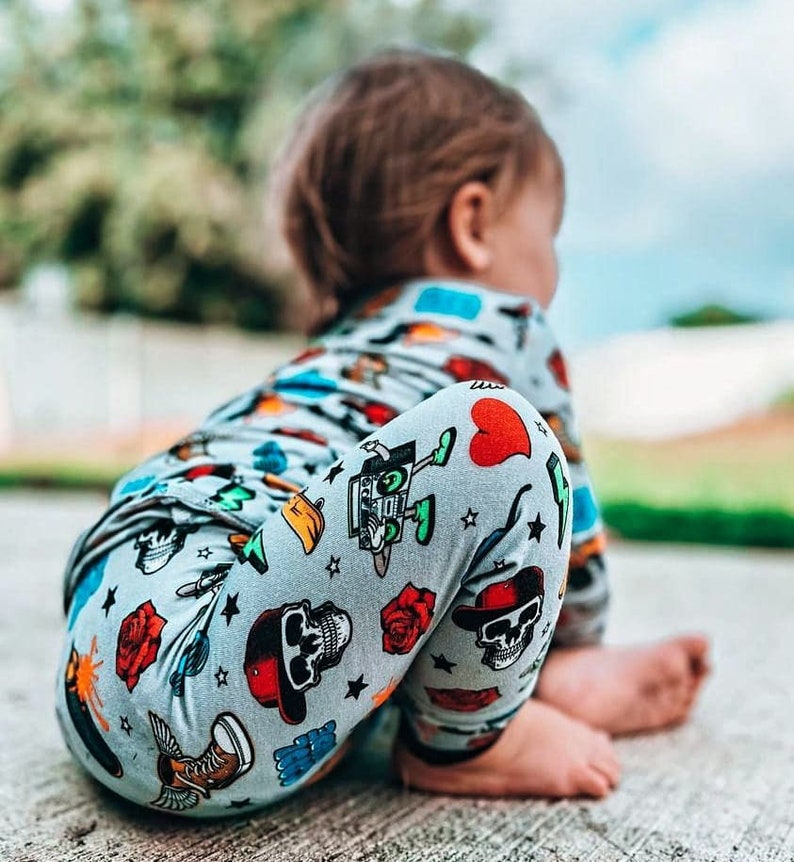 May include: A child wearing a light gray onesie and leggings with a repeating pattern of skulls, roses, hearts, and other graphics. The child is sitting on a concrete surface, with their back to the camera.