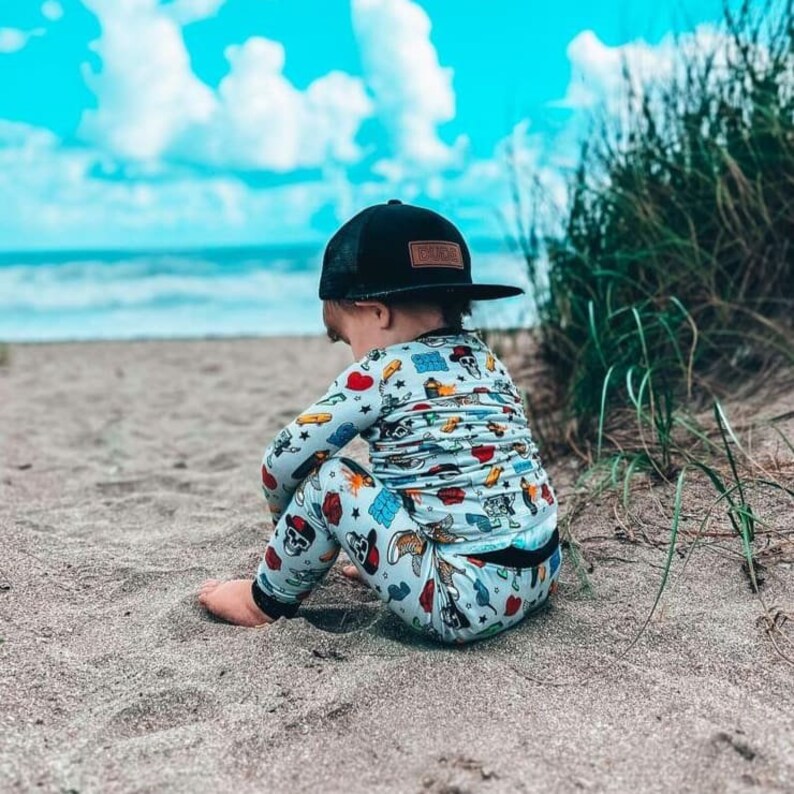 May include: A child wearing a black trucker hat with a brown patch and a light blue pajama set with a colorful print of skulls, hearts, and other graphics. The child is sitting on the sand near the ocean.
