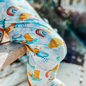 May include: A light blue and white patterned onesie with a rainbow, rain boots, and umbrella design. The onesie has a checkered pattern and is worn by a child. The background is blurred, with trees and a bench visible.
