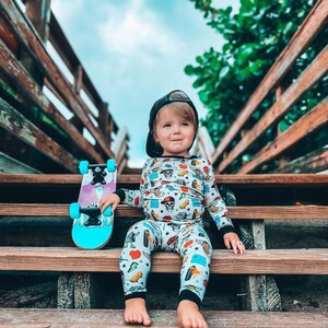 May include: A child wearing a black cap and patterned pajamas sits on wooden steps, holding a teal and purple skateboard. The pajamas feature various colorful illustrations. The background includes a wooden railing and a cloudy sky.