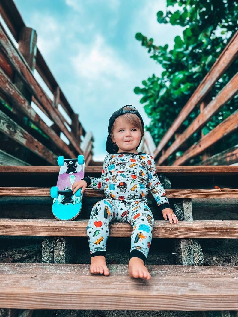 May include: A child wearing a black cap and patterned pajamas sits on wooden steps, holding a teal and purple skateboard. The pajamas feature various colorful illustrations. The background includes a wooden railing and a cloudy sky.
