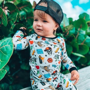 May include: A child wearing a gray pajama set with a colorful print of skulls, sneakers, hearts, and the words "Cool Dude." The child is also wearing a black mesh trucker hat. The background features green foliage and a blue sky.