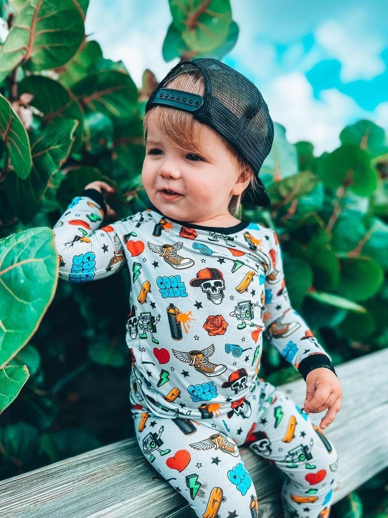 May include: A child wearing a gray pajama set with a colorful print of skulls, sneakers, hearts, and the words "Cool Dude." The child is also wearing a black mesh trucker hat. The background features green foliage and a blue sky.