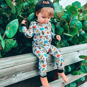 May include: A child wearing a light gray pajama set with a colorful pattern of hearts, lightning bolts, skateboards, and skulls. The child is also wearing a black baseball cap and is sitting on a wooden fence.