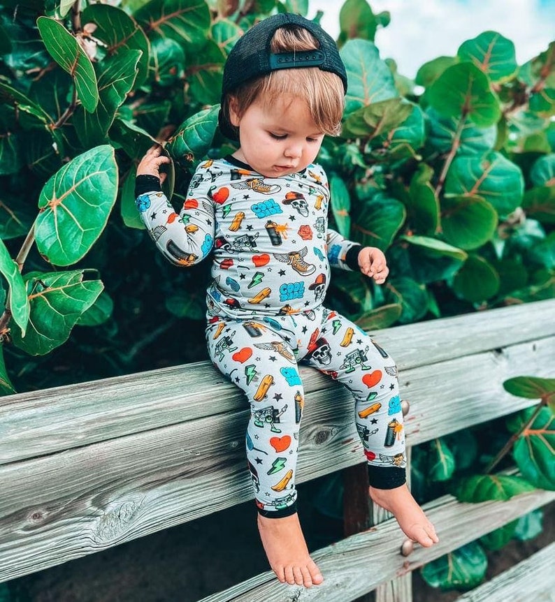 May include: A child wearing a light gray pajama set with a colorful pattern of hearts, lightning bolts, skateboards, and skulls. The child is also wearing a black baseball cap and is sitting on a wooden fence.