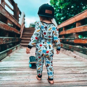 May include: A child wearing a black baseball cap with "DUDE" on a brown patch, and a light blue pajama set with colorful graphics. The child is standing on a wooden bridge with a skateboard.