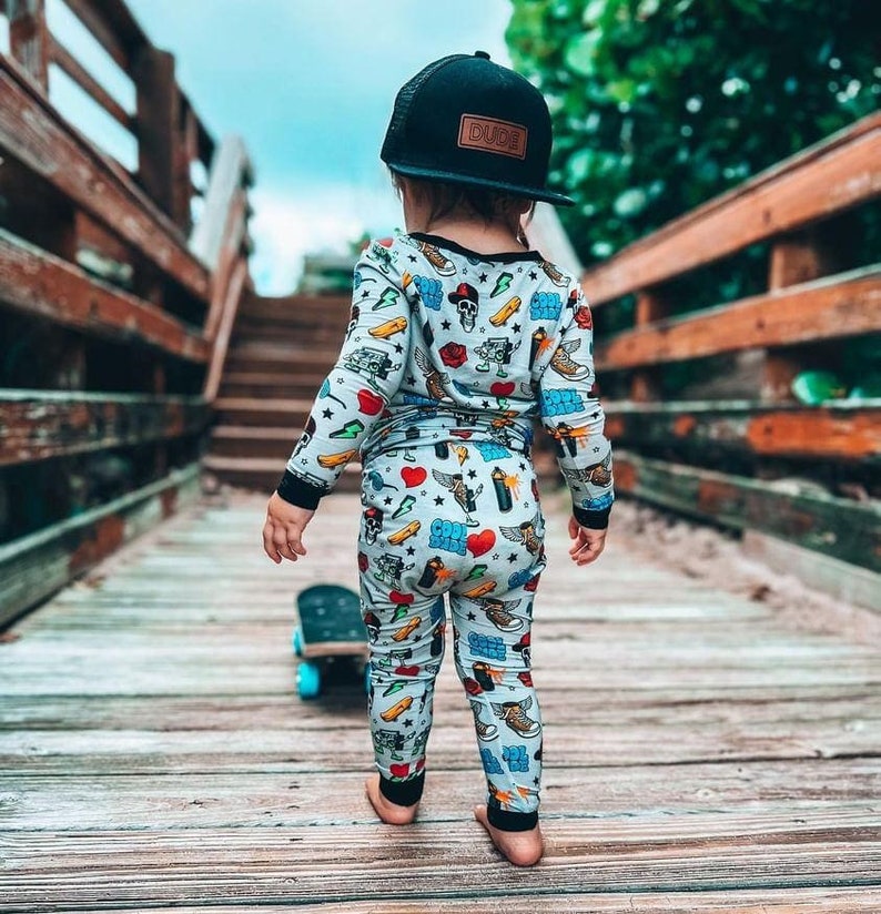 May include: A child wearing a black baseball cap with "DUDE" on a brown patch, and a light blue pajama set with colorful graphics. The child is standing on a wooden bridge with a skateboard.