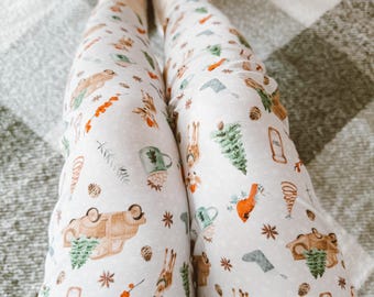 Puede incluir: Leggings blancos con un estampado festivo que incluye renos, pinos, adornos y un cardenal rojo. El estampado es en tonos de verde, marrón, rojo y blanco.