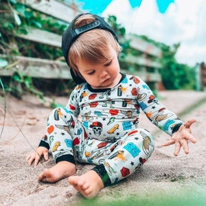 May include: A child wearing a black baseball cap and a light gray pajama set with a colorful print of skulls, roses, skateboards, and other graphics. The child is sitting on the sand.