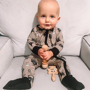 May include: A baby wearing a brown and black dinosaur print onesie is sitting on a white chair. The baby is holding a wooden rattle in one hand and a wooden teether in the other hand.