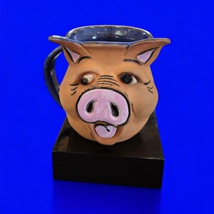 May include: A ceramic mug shaped like a pig head with a tan face, a prominent pink snout, and dark blue accents on the handle and interior rim. The pig features sculpted ears, dark eyes, and a small tongue visible at the bottom of the snout.