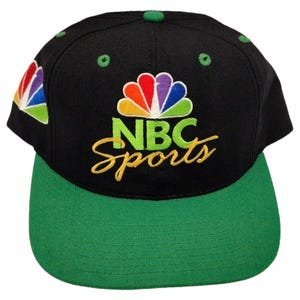 May include: A black baseball cap with a green brim and green eyelets. The front features the embroidered NBC peacock logo in rainbow colors above the text NBC Sports in lime green and gold script. A smaller NBC peacock logo is on the side.