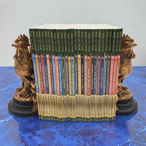 May include: A collection of "Magic Tree House" books, held between two bronze-colored rooster bookends. The book spines display titles and author's name. The bookends sit on a blue surface.