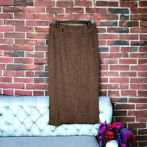 Peut inclure: Une jupe mi-longue marron à la texture chinée, dotée d'une coupe droite et d'une taille haute. Le tissu présente un motif moucheté subtil sur toute la surface. La jupe est présentée sur un cintre noir devant un mur en briques rouges.
