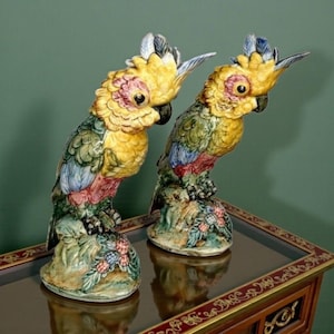 Peut inclure: Deux figurines de perroquets en céramique. Les perroquets ont des crêtes jaunes, des plumes multicolores et sont perchés sur une base avec des détails floraux et de baies. Les figurines sont exposées sur une table en bois foncé avec une bordure décorative.