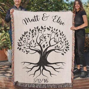 Op de afbeelding: Een wit geweven wandkleed met een zwart boomontwerp en de tekst "Matt & Elise" en "5/12/16". Twee mensen houden het wandkleed vast.