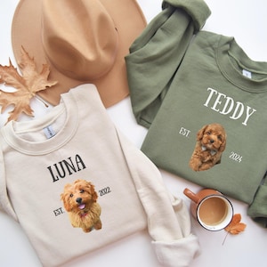 Op de afbeelding: Twee crèmekleurige en olijfgroene sweatshirts met hondenportretten en gepersonaliseerde tekst. Het crèmekleurige sweatshirt heeft een bruine hond met de tekst "LUNA EST. 2022". Het olijfgroene sweatshirt heeft een bruine hond met de tekst "TEDDY EST. 2024".