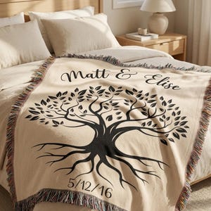 May include: A cream-coloured woven throw blanket featuring a black graphic of a tree with roots. The text "Matt & Elise" is printed in a script font above the tree, and the date "5/12/16" is printed in a sans-serif font below the roots.
