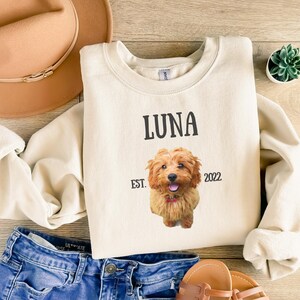May include: A cream-colored sweatshirt with the name "LUNA" printed in black, along with the text "EST. 2022". A photo of a brown and white dog is printed on the sweatshirt.