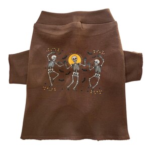 May include: A brown dog shirt featuring a graphic of three dancing skeletons with a yellow sun in the center. The skeletons are white, and the shirt has long sleeves. The design includes bats and autumn leaves.