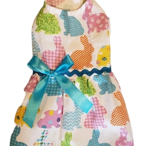 May include: A white pet dress featuring a colorful bunny pattern in various colors and textures. The dress has a blue ribbon bow and trim. The overall design is festive and suitable for Easter or spring.