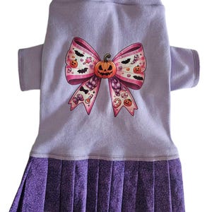 May include: A lavender dog dress with a Halloween-themed bow design. The bow features a jack-o'-lantern, bats, and candy. The dress has a pleated skirt in a darker purple fabric.