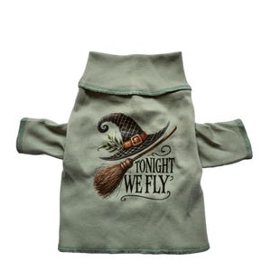 May include: Sage green pet shirt with a witch hat and broom graphic. The text "TONIGHT WE FLY" is printed below the graphic. The shirt has a high collar and sleeves with green trim.