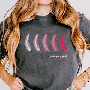May include: A dark gray t-shirt with a graphic of four pink feathers and the text "finding my pink".