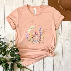 May include: A peach-colored t-shirt with a floral design inside the outline of the state of Wisconsin.