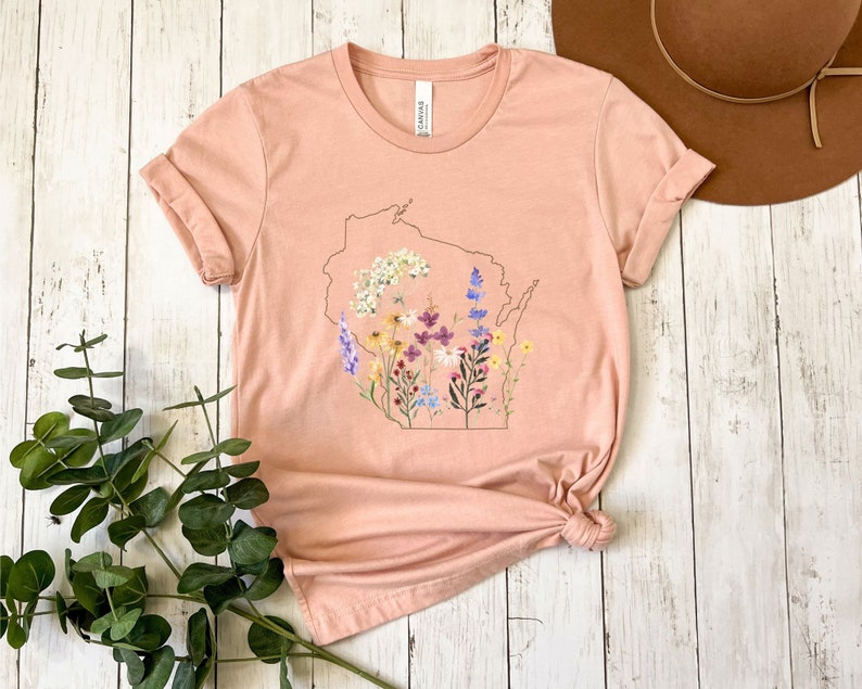 May include: A peach-colored t-shirt with a floral design inside the outline of the state of Wisconsin.