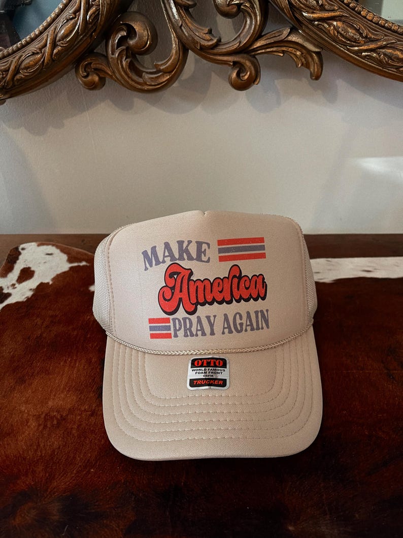 Peut inclure: Un chapeau de camionneur beige avec un motif de drapeau am&eacute;ricain rouge, blanc et bleu. Le chapeau dit "Make America Pray Again" en lettres rouges et blanches. Le chapeau a un dos en maille noire et une &eacute;tiquette blanche qui dit "Otto World Famous Foam Front Trucker".