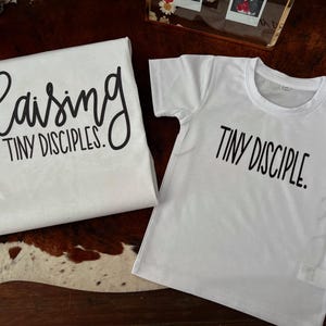 May include: Two white t-shirts with black text. One shirt reads "Raising TINY DISCIPLES." in a script font. The other shirt reads "TINY DISCIPLE." in a sans-serif font. The shirts are displayed on a brown and white patterned surface.