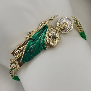 May include: A green and gold bracelet with a charm of Saint Martin, a religious figure holding a flag and a staff. The charm is surrounded by a halo and has a white background.
