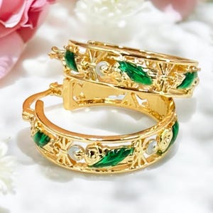 May include: A pair of gold-colored hoop earrings with intricate detailing. The earrings feature green enamel accents and small, round, white embellishments. The design includes floral and geometric patterns. The earrings are displayed on a white surface with pink and white flowers in the background.