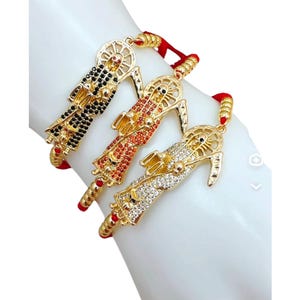 May include: Three red string bracelets with gold charms featuring a skeleton figure with a scythe. The charms are adorned with black, red, and clear rhinestones.