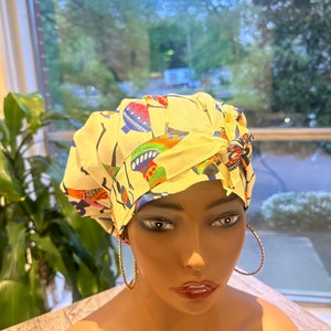 Op de afbeelding: Een crèmekleurige hoofddoek met een kleurrijk patroon van abstracte vormen en figuren. De hoofddoek is aan de zijkant geknoopt en versierd met een decoratieve speld. Grote gouden oorringen maken de look compleet.